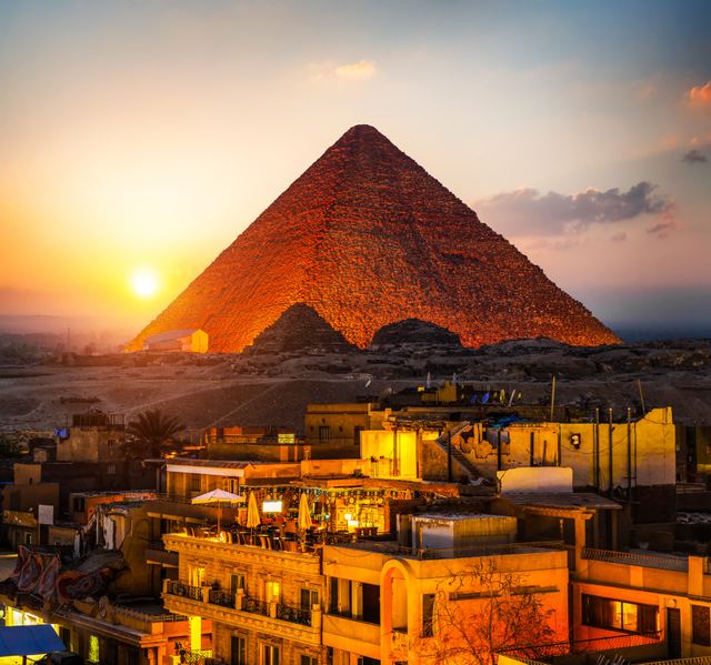A pyramid at Giza glowing orange at sunset, above the rooftops of the city in the foreground.