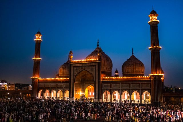 Jama Masjid in Delhi lit up at dusk, with its domes and two tall minarets above a large crowd.