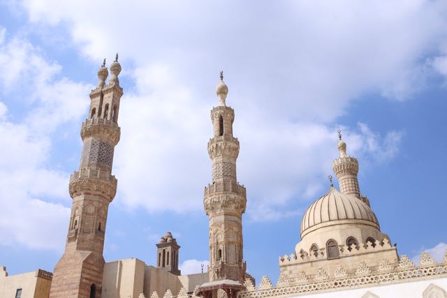 Two carved stone minarets and a ribbed white dome of Al-Azhar Mosque in Cairo against a blue sky with clouds.