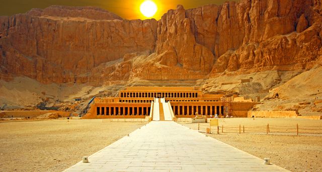 The terraced colonnades of the Temple of Hatshepsut against tall cliffs, with a long ramp leading up and the sun low above the cliff.