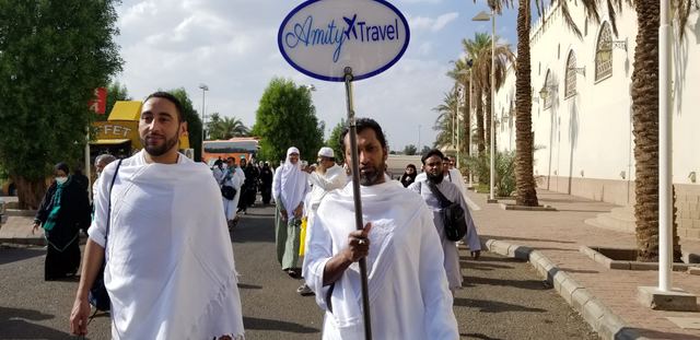 Amity group leader in ihram holding the Amity Travel sign, leading pilgrims along a palm-lined road