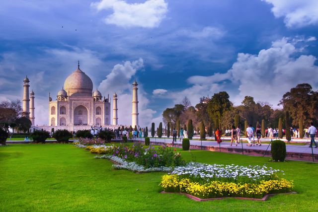 The Taj Mahal in Agra beyond gardens with lawns and flowerbeds under a cloudy blue sky.