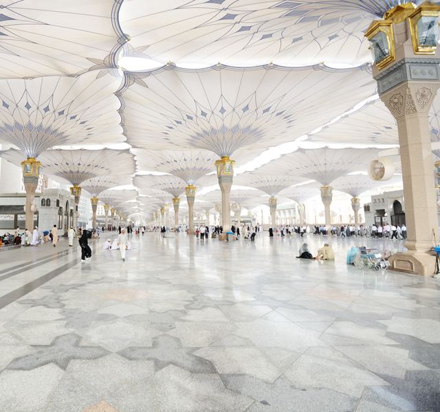 Visitors walking across the marble courtyard of Masjid an-Nabawi in Madinah beneath rows of open white umbrellas.