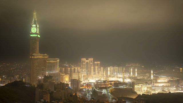 The Makkah clock tower lit green at night above the glowing city and Masjid al-Haram.