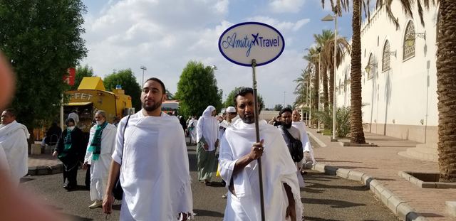Amity group leader in ihram holding the Amity Travel sign, leading pilgrims along a palm-lined road