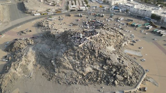 Aerial view of Jabal al-Rahmah at Arafat, with visitors on the rocky hill and tour buses parked around it.