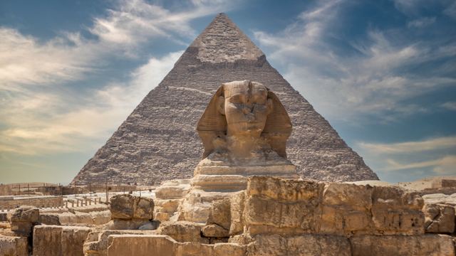 The Great Sphinx in front of a pyramid at Giza under a blue sky with clouds.