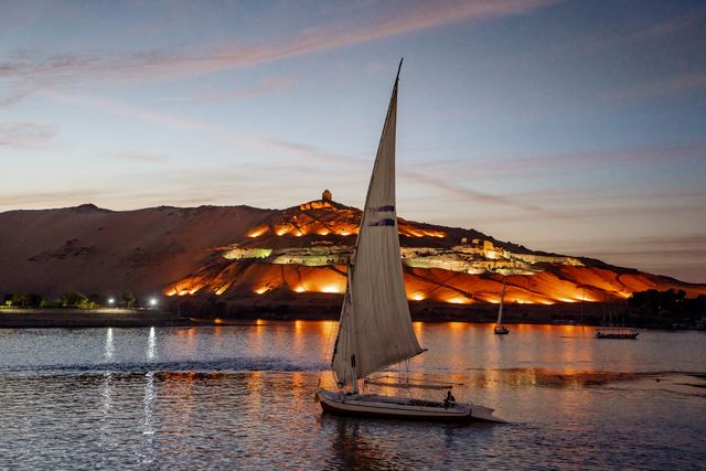 A felucca sailboat on the Nile at dusk, with lights along the hillside on the far bank.