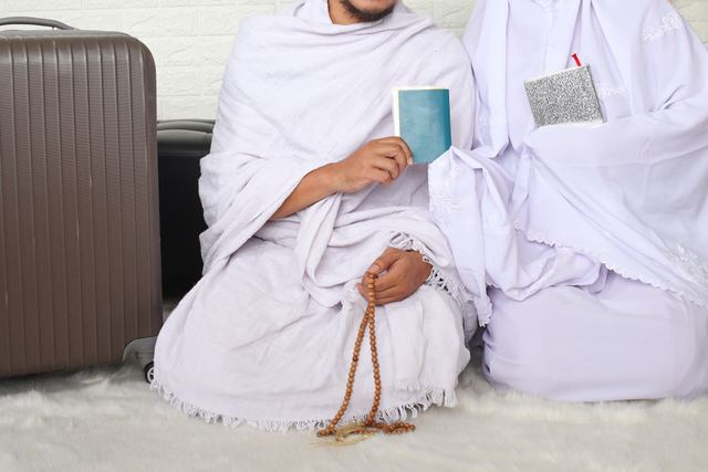 Two people in white ihram clothing seated beside suitcases, holding small books and prayer beads.
