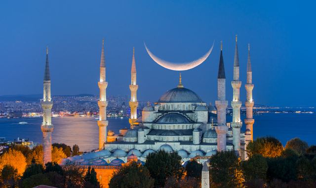 The Blue Mosque in Istanbul at dusk, with its domes and minarets, a crescent moon above and the sea behind.