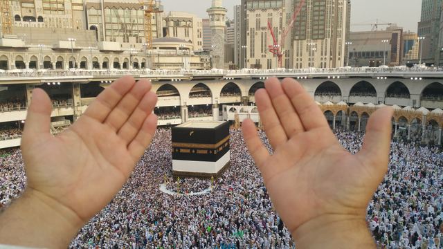 Two open hands raised toward the Ka‘bah and the crowded courtyard of Masjid al-Haram.