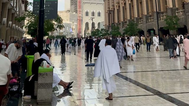 Pilgrims, some in white ihram, walk along a wide marble plaza towards Masjid al-Haram in Makkah.