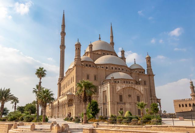 The Mosque of Muhammad Ali in Cairo, with its domes and two slender minarets, palm trees in front and a blue sky.