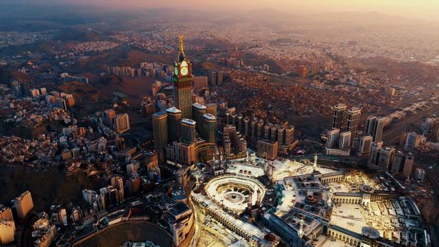 Aerial view of Makkah at golden hour: Masjid al-Haram glowing in warm light, the clock tower rising beside it and the city stretching to a hazy horizon.