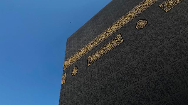 Close view of the Kaaba's black kiswah and its gold calligraphy band against a clear blue sky in Makkah.